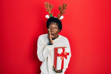 Geyik Noel şapkası takan genç Afrikalı Amerikalı adam elinde hediyeyle korkmuş ve şok olmuş, şaşırmış ve hayrete düşmüş bir yüz ifadesi takınıyor. 