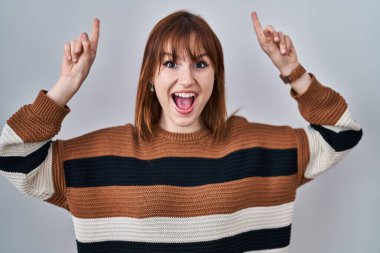 İzole edilmiş arka planda çizgili süveter giyen genç ve güzel bir kadın. Şaşırmış, şaşırmış, parmaklarıyla işaret ediyor ve kollarını kaldırıyordu.. 