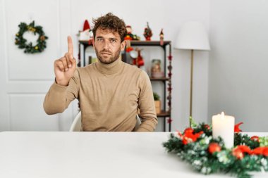 Noel süslemesi masasında oturan sakallı yakışıklı genç adam parmağını kaldırmış ve kızgın bir ifadeyle işaret ediyor, hiçbir hareket göstermiyor. 