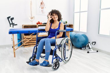 Young middle eastern woman sitting on wheelchair at physiotherapy clinic smiling positive doing ok sign with hand and fingers. successful expression. 