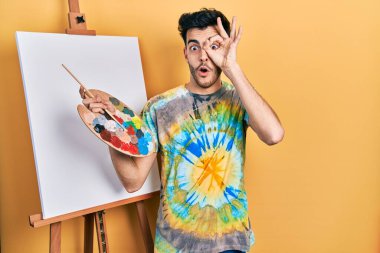 İspanyol asıllı genç adam paletle ayakta dikiliyor, resim yapıyor, şaşırmış yüz ifadesiyle şaşırmış, parmaklarının arasından bakıyor. İnançsız ifade. 