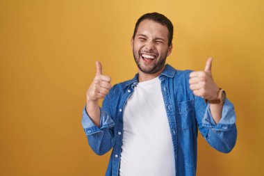 Hispanic man standing over yellow background success sign doing positive gesture with hand, thumbs up smiling and happy. cheerful expression and winner gesture. 