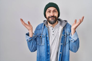 Young hispanic man with tattoos wearing wool cap clueless and confused expression with arms and hands raised. doubt concept. 