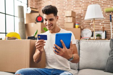 Young hispanic man using touchpad and credit card sitting on sofa at new home