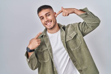 Soyutlanmış arka planda duran, neşeli gülümseyen ve parmaklarıyla ve ağzıyla işaret eden İspanyol genç bir adam. diş sağlığı kavramı. 