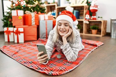 Kafkasyalı genç bir kız Noel şapkası takıyor ve akıllı telefonuyla evde yerde selfie çekiyor..