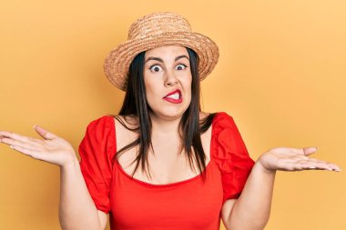 Yazlık şapkalı genç İspanyol kadın elleri ve kolları havada, kafası karışmış bir ifade takınıyor. şüphe kavramı. 