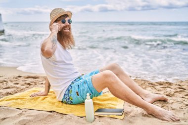 Genç kızıl saçlı adam kumsalda havlunun üzerinde oturmuş akıllı telefondan konuşuyor..