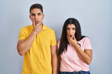 İzole edilmiş arka planda duran genç çift, elleri çenesinde olan inançsızlıktan, şaşkınlıktan ve şaşkınlıktan büyülenmiş görünüyor. 