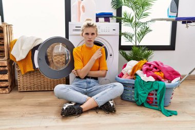 Young blonde woman doing laundry sitting by washing machine pointing with hand finger to the side showing advertisement, serious and calm face 