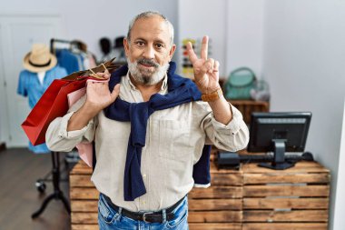 Butik dükkanında alışveriş torbaları tutan yakışıklı bir adam kameraya gülümseyen bir gülümsemeyle parmaklarıyla zafer işareti yapıyor. İki numara.. 