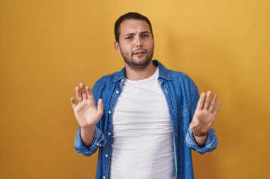 Hispanic man standing over yellow background moving away hands palms showing refusal and denial with afraid and disgusting expression. stop and forbidden. 