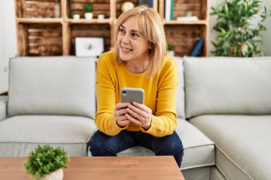 Orta yaşlı sarışın kadın akıllı telefon kullanıyor. Evdeki kanepede oturuyor..