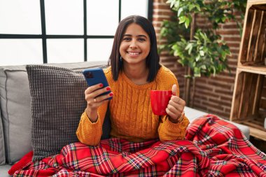Genç Latin kadın kahve içiyor ve evde akıllı telefon kullanıyor.