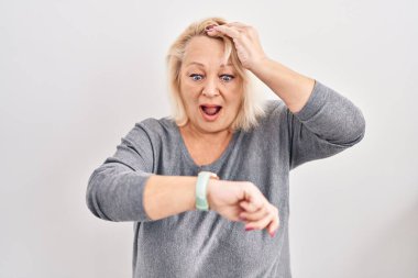Middle age caucasian woman standing over white background looking at the watch time worried, afraid of getting late 
