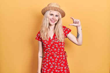 Beautiful caucasian woman with blond hair wearing summer hat smiling and confident gesturing with hand doing small size sign with fingers looking and the camera. measure concept. 