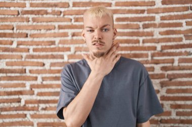 Young caucasian man standing over bricks wall cutting throat with hand as knife, threaten aggression with furious violence 