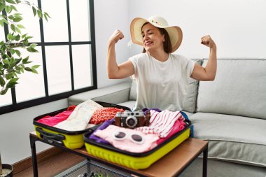 Beautiful middle age hispanic woman packing summer clothes in suitcase showing arms muscles smiling proud. fitness concept. 