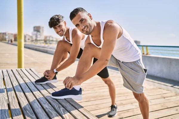 İki İspanyol erkek, deniz kenarında bağcıklarını bağlayarak gülümsüyor.