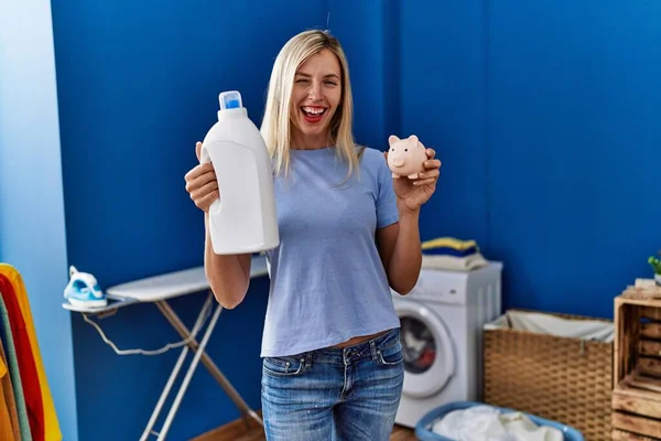 Beautiful woman doing laundry holding detergent bottle and piggy bank winking looking at the camera with sexy expression, cheerful and happy face. 