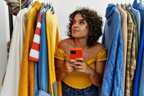 Young latin customer woman smiling happy appearing through clothes using smartphone at clothing store.