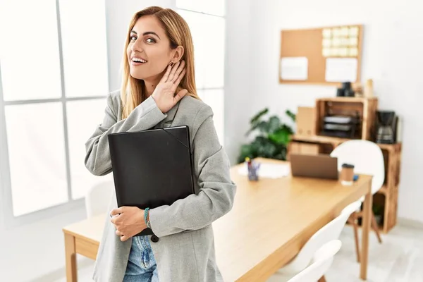 Blonde business woman at the office smiling with hand over ear listening an hearing to rumor or gossip. deafness concept.