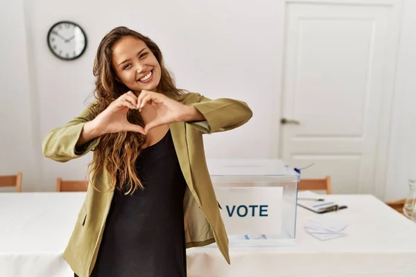 Beautiful hispanic woman standing at political campaign room smiling in love doing heart symbol shape with hands. romantic concept.