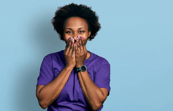 African american woman with afro hair wearing casual purple t shirt laughing and embarrassed giggle covering mouth with hands, gossip and scandal concept