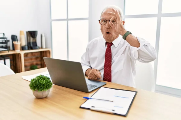 Senior man working at the office using computer laptop doing ok gesture shocked with surprised face, eye looking through fingers. unbelieving expression.