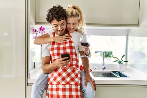 若いカップル幸せな抱擁笑みを浮かべてカウンターに座っている 赤ワインを飲み キッチンでスマートフォンを使う — ストック写真