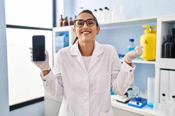Young hispanic woman working at scientist laboratory with smartphone screaming proud, celebrating victory and success very excited with raised arm