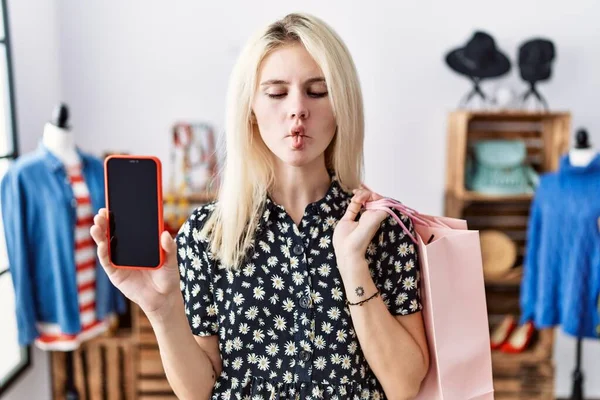 Young blonde woman holding shopping bags showing smartphone screen making fish face with mouth and squinting eyes, crazy and comical.