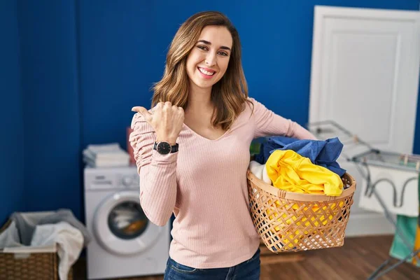 Young woman holding laundry basket pointing to the back behind with hand and thumbs up, smiling confident