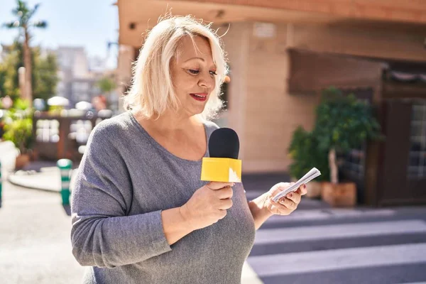 Middle age blonde woman reporter working using microphone at street