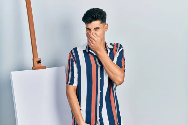 Young Hispanic Man Standing Close Empty Canvas Smelling Something Stinky — Foto de Stock