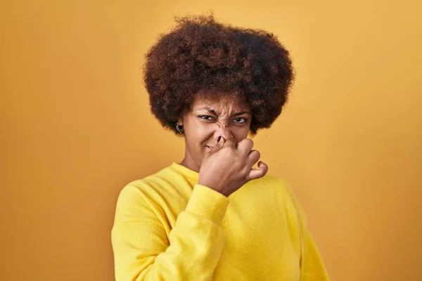 年轻的非洲裔美国女人站在黄色的背景上 闻到一股难闻难闻的气味 用手指捂住鼻子呼吸 — 图库照片