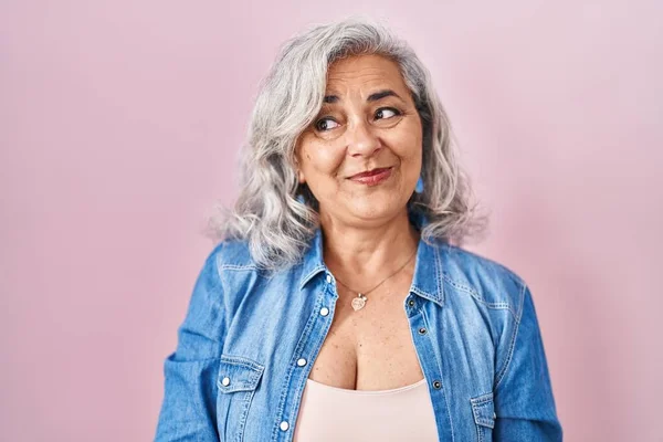 Middle age woman with grey hair standing over pink background smiling looking to the side and staring away thinking.