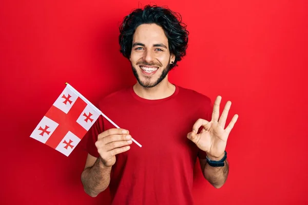 ジョージアの旗を持っているハンサムなヒスパニック系の男は指でOkサインをします 笑顔優しいジェスチャー優れたシンボル — ストック写真