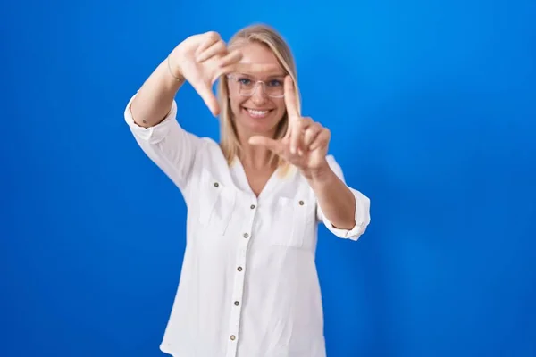 青い背景に立っている若い白人女性は 幸せな顔をした手と指でフレームを作り笑顔 創造性と写真の概念 — ストック写真