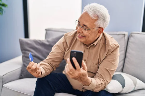 Senior man using smartphone and credit card sitting on sofa at home