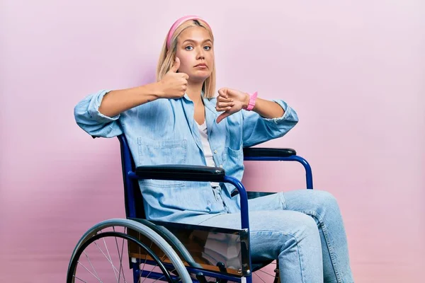 Beautiful blonde woman sitting on wheelchair doing thumbs up and down, disagreement and agreement expression. crazy conflict