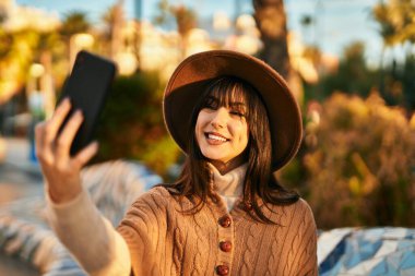 Kışlık şapkalı esmer kadın parkta akıllı telefonuyla selfie çekiyor.