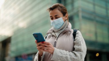 Orta yaşlı, tıbbi maske takan bir kadın sokakta akıllı telefon kullanıyor.