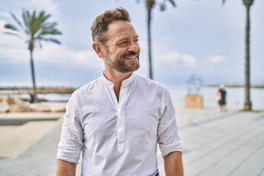 Orta yaşlı bir adam deniz kenarında kendinden emin gülümsüyor.