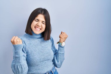 Young hispanic woman standing over blue background very happy and excited doing winner gesture with arms raised, smiling and screaming for success. celebration concept. 
