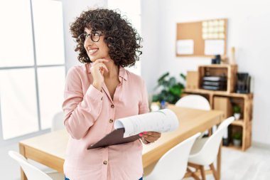 Young middle eastern woman wearing business style at office with hand on chin thinking about question, pensive expression. smiling and thoughtful face. doubt concept. 