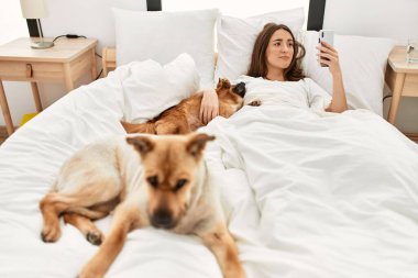 Genç İspanyol kadın köpeğe sarılıyor ve yatakta akıllı telefon kullanıyor.