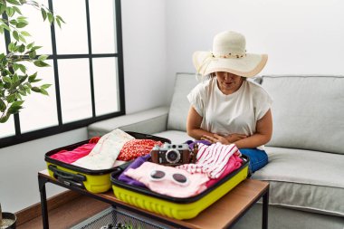 Beautiful middle age hispanic woman packing summer clothes in suitcase with hand on stomach because nausea, painful disease feeling unwell. ache concept. 