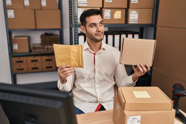 Young hispanic man with beard working at small business ecommerce holding delivery packages smiling looking to the side and staring away thinking. 