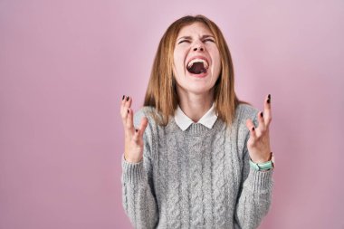 Beautiful woman standing over pink background crazy and mad shouting and yelling with aggressive expression and arms raised. frustration concept. 